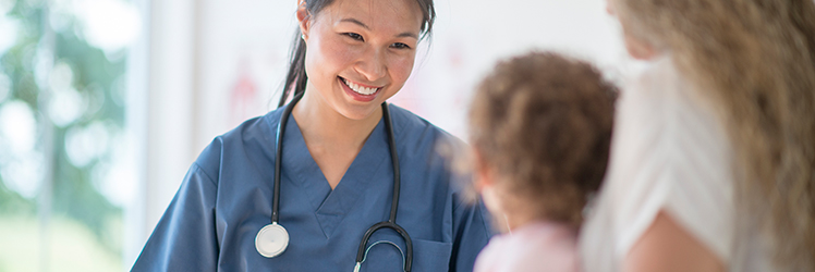 A nurse in scrubs smiles at a mother and her young daughter.