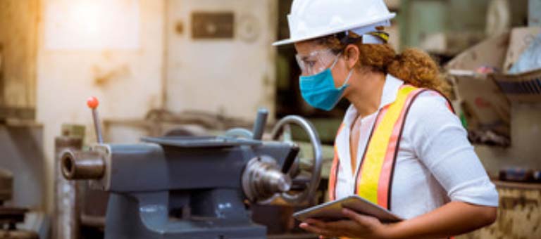 Set inside a workshop, a woman, walking in profile, wears personal protective equipment (PPE), including a blue disposable N95 mask, a white hardhat, a high-visibility safety vest and safety glasses