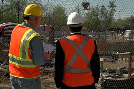 Set on a construction jobsite, two men wearing personal protective equipment, including hardhats and high-visibility safety vests, stand with their backs to the camera observing the jobsite.