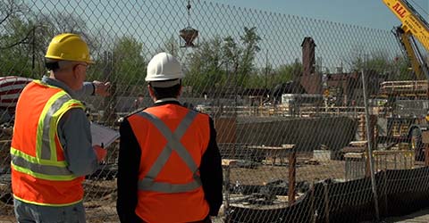 Set on a construction jobsite, two men wearing personal protective equipment, including hardhats and high-visibility safety vests, stand with their backs to the camera observing the jobsite.