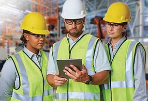 Set in an industrial facility, three people, two women and a man, stand together looking at a tablet computer. All wear personal protective equipment (PPE), including hardhats, safety glasses and high-visibility safety vest.