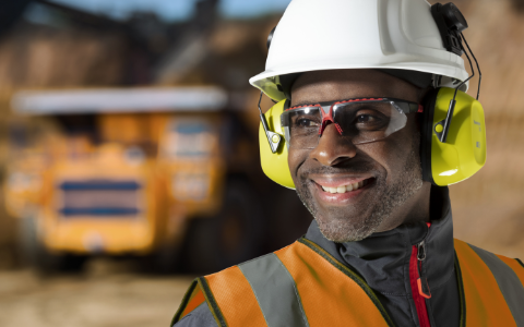 A worker wearing Honeywell Uvex safety glasses, protective earmuffs and a hardhat smiles on a jobsite.