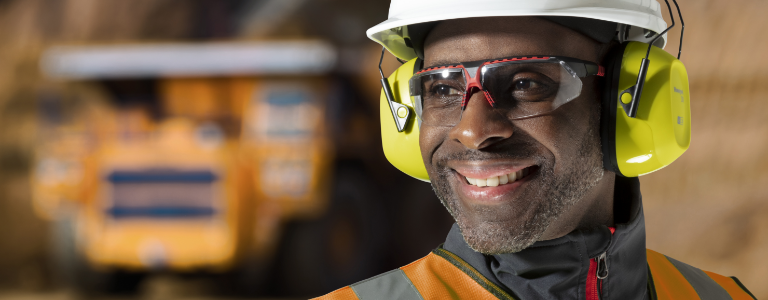 A worker wearing Honeywell Uvex safety glasses, protective earmuffs and a hardhat smiles on a jobsite.