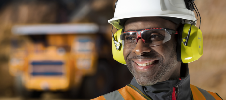 A worker wearing Honeywell Uvex safety glasses, earmuffs and a hardhat smiles on a jobsite.