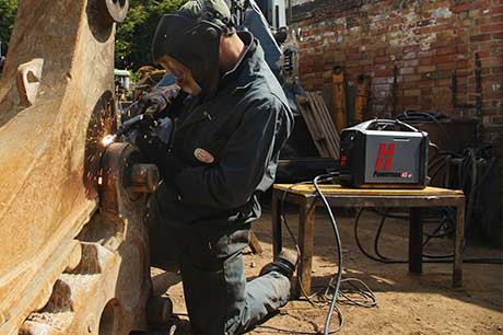 While kneeling, a man wearing welding safety helmet, welding jacket and welding gloves, uses a Hypertherm Powermax plasma-cutting system on a large machine part outside in the workyard.