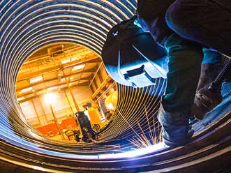 Welder standing inside of a large corrugated metal pipe performing a weld on the interior of the pipe underneath.