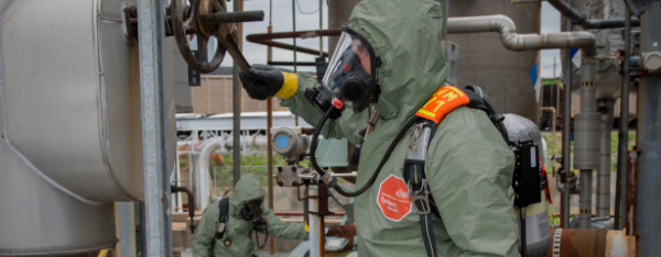 Worker is completely covered by DuPont protective clothing as he works outside next to machines, tanks and gears.