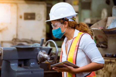 Set inside a workshop, a woman, walking in profile, wears personal protective equipment (PPE), including a blue disposable N95 mask, a white hardhat, a high-visibility safety vest and safety glasses.