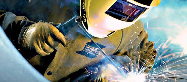 A welder performs a stick-welding application while wearing ESAB branded welding safety clothing and personal protective equipment (PPE), including a welding helmet, welding gloves and a welding jacket.